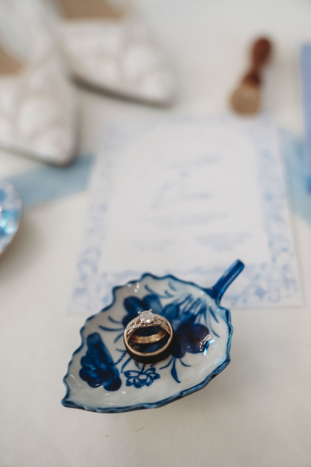 Gold wedding rings on a blue and white floral leaf-shaped dish, with bridal shoes and invitation in the background.