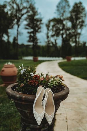 White lace bridal heels resting on a floral garden urn along a scenic wedding venue pathway.