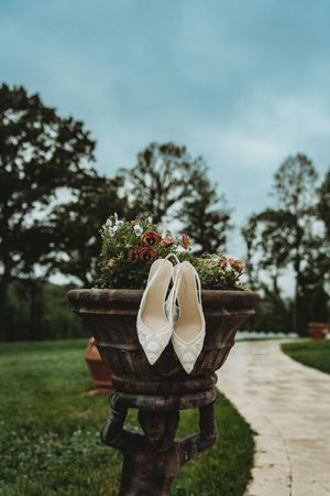 White lace bridal heels resting in a rustic stone planter with colorful flowers, set along a garden pathway.