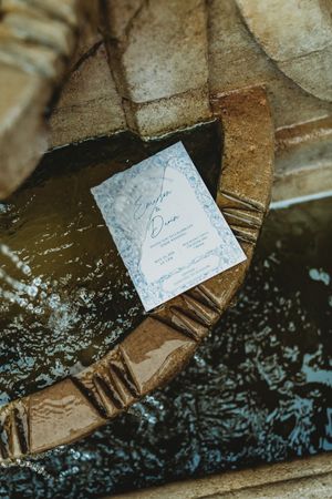 Elegant wedding invitation resting on a stone fountain ledge with blue floral details and script typography.