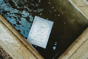 Wedding invitation card resting on the edge of a pool with blue water, featuring elegant blue typography.
