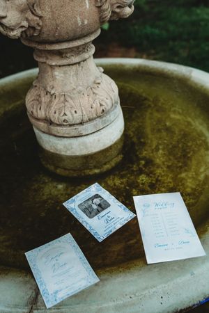 Wedding stationery suite displayed in a stone fountain, featuring programs and cards with blue calligraphy details.