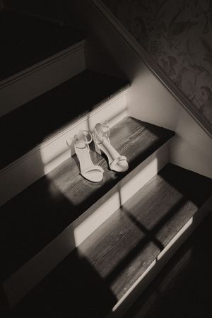 Black and white photo of elegant high heel shoes resting on wooden staircase steps in soft natural light.
