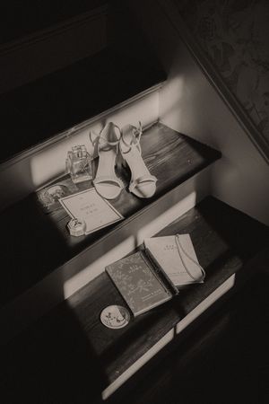 Black and white photo of bridal accessories on wooden stairs, featuring heels, perfume, books, and a compact mirror.