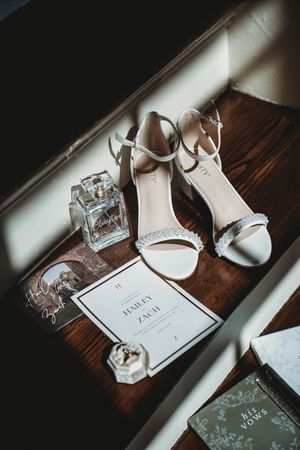 White bridal heels, perfume, wedding invitation for Hailey and Zach, and a vow book arranged on a wooden surface.