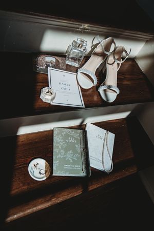 Elegant wedding flat lay featuring white heels, vow books, invitations, perfume, and jewelry on wooden stairs in soft natural light.