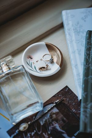 Wedding rings and jewelry displayed on a ceramic dish with perfume bottle and stationery on a wooden surface.