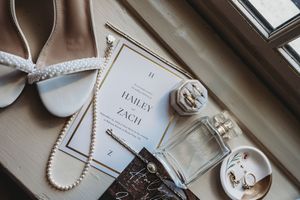 Elegant wedding flat lay featuring bridal shoes, pearl necklace, perfume, rings, and a Hailey and Zach wedding invitation.