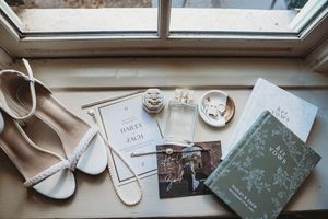 Flat lay of wedding details including heels, invitations, perfume, rings, and a green vow book on a wooden windowsill.
