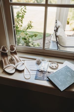 Bridal flat lay with white heels, pearl necklace, wedding invitations, and a wish book on a sunlit windowsill.