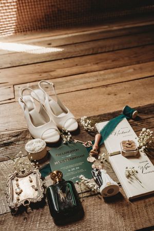 Elegant wedding flat lay with white heels, green invitation, perfume, floral accents, ring box, and vintage accessories on wood floor.
