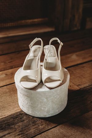White bridal slingback heels displayed on a decorative round box on rustic wooden floor.