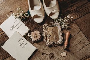 Elegant wedding flat lay with white heels, gold rings, earrings, wax seal, baby's breath, and vow books on rustic wood.