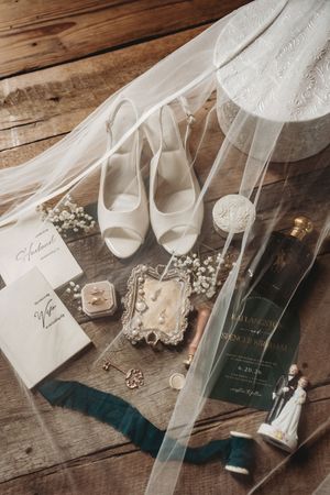 Elegant bridal flat lay with white heels, wedding rings, invitations, perfume, and accessories on rustic wood under a veil.