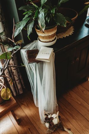 Wedding vows booklet and veil draped over dark wooden dresser with potted plants and floral bouquet.