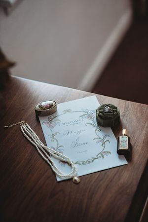 Wedding flat lay with floral program, pearl necklace, ring box, vintage perfume bottle, and decorative trinket box on wood table.