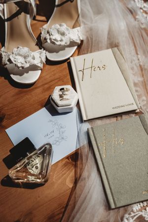 Wedding flat lay with lace heels, His and Hers vow books, diamond ring box, perfume bottle, and floral stationery on wood.