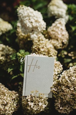 Gray "His" vow book resting among white hydrangea blooms in a lush garden setting.