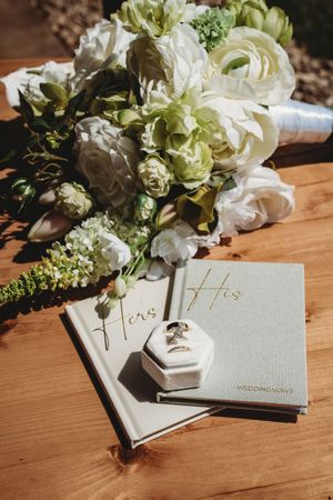 Wedding vow books labeled His and Hers with diamond rings in a white box beside a white and green bridal bouquet.