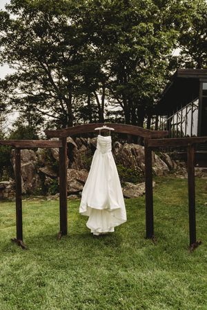 White wedding dress hanging on a rustic wooden arch in a lush green garden with stone walls and trees in the background.
