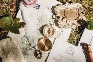 Vintage travel flat lay with brass compass, antique tray, maps, and elegant stationery on green grass.