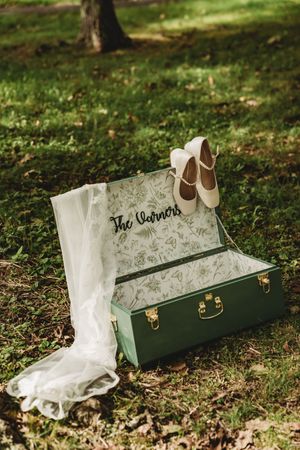 Green vintage suitcase with floral lining, The Corners sign, bridal veil, and ivory wedding shoes on grass.