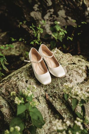 White bridal heels with crystal ankle straps resting on a moss-covered rock surrounded by lush green foliage.