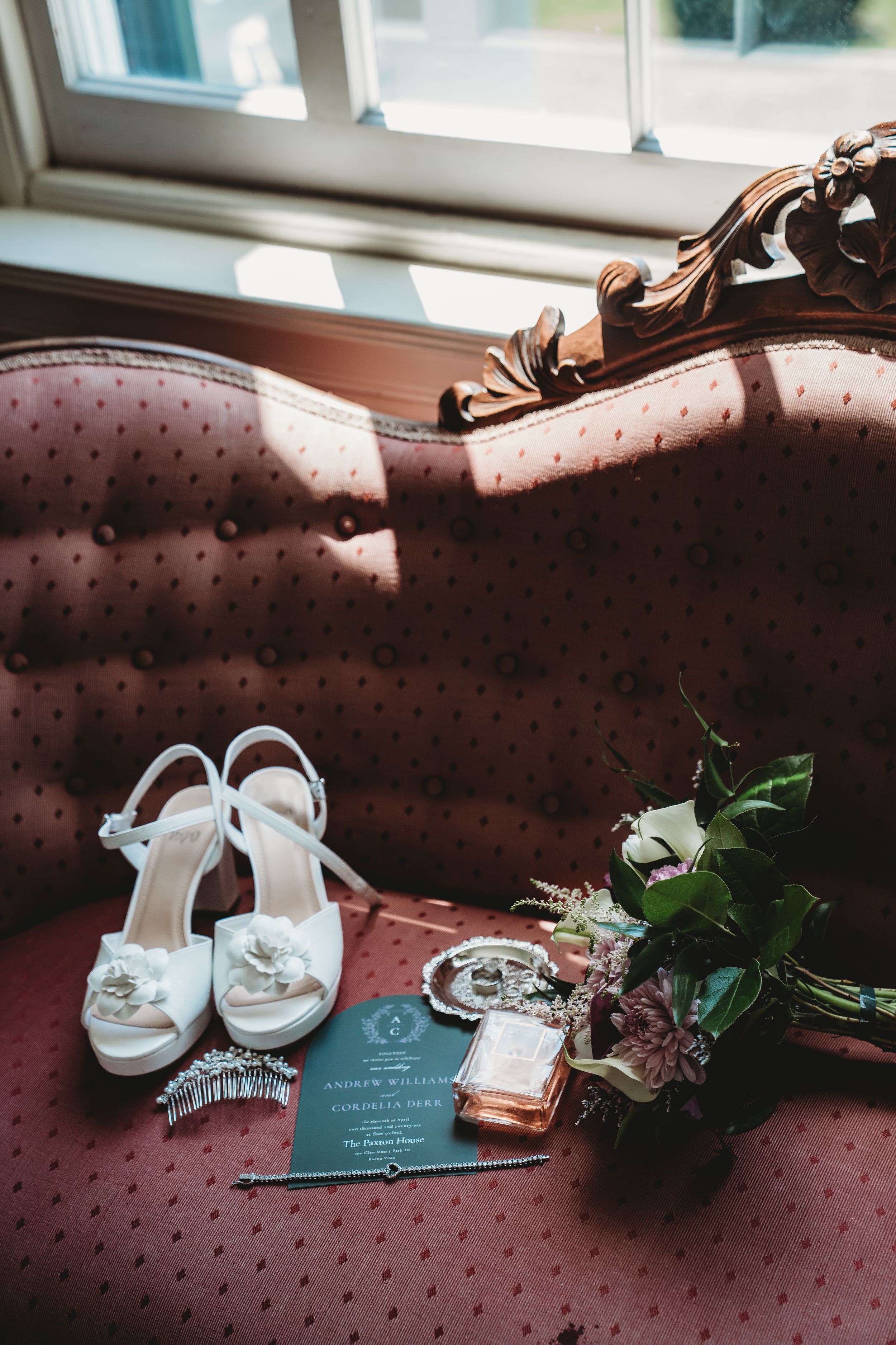 Elegant wedding flat lay with white heels, floral bouquet, invitation, pearl jewelry, and perfume on a tufted pink chair.
