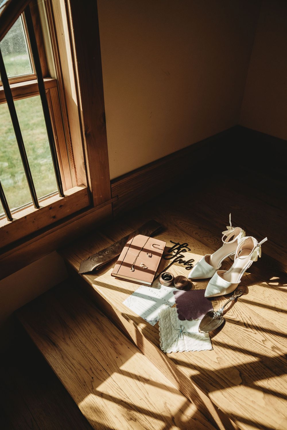 Wedding accessories including shoes and jewelry sit in dramatic window light on a wooden floor.