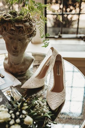 Elegant white bridal shoes with pearl embellishments displayed next to stone planters with delicate greenery.