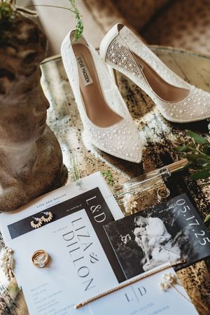 Wedding details featuring elegant white bridal shoes, wedding rings, and black invitation cards arranged artistically.
