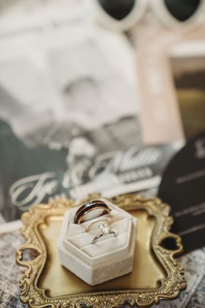 Gold ornate tray with diamond engagement ring in velvet box and wedding band, with vintage photos in background.