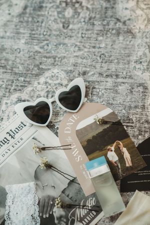 Heart-shaped sunglasses rest on a save-the-date card with couple photo, bobby pins, and lace on a gray textured surface.