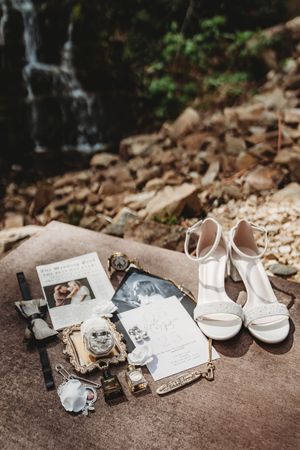 White bridal heels and wedding stationery flat lay with rings and accessories on rocks near a waterfall.