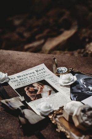 Wedding Post newspaper invitation with couple photo, white roses, pocket watch, and mirror on stone surface.