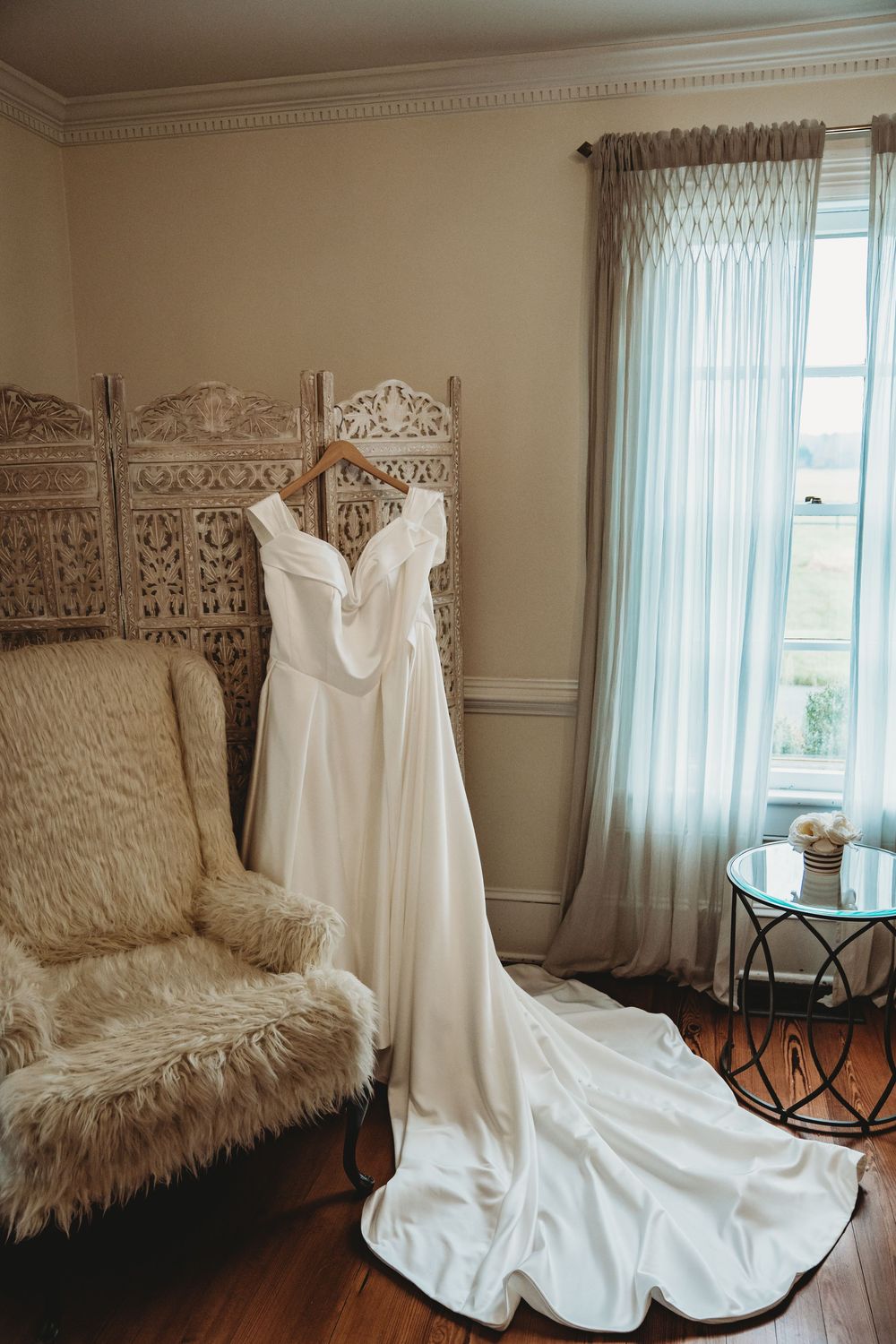 Elegant white wedding gown with train hanging on wooden hanger in a bright bridal suite with ornate screen.