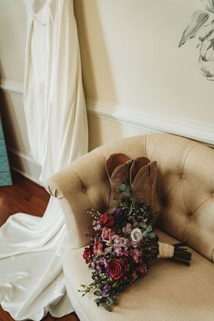 Bridal bouquet of red, pink, and purple flowers with cowboy boots on a tufted beige chair beside a white wedding dress.