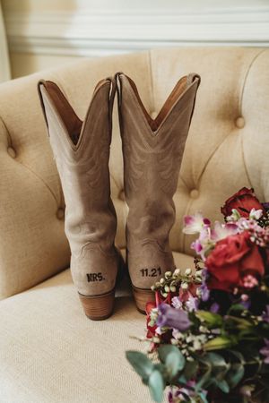 Personalized cowboy boots with MRS. and 11.21 wedding date beside a bridal bouquet on a tufted chair.