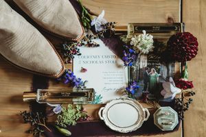 Wedding flat lay with invitation, floral accents, perfume bottles, rings, and beige shoes on a wooden surface.