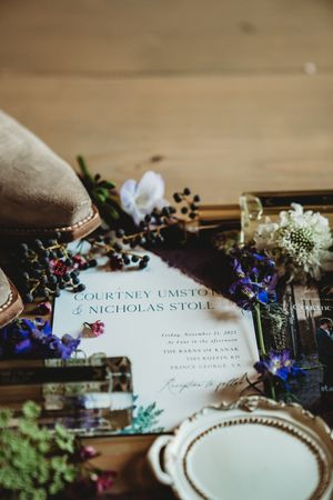 Wedding invitation for Courtney Umstot and Nicholas Stoll surrounded by wildflowers, berries, and gold accessories on wood.