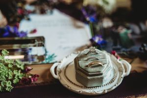 Wedding rings displayed on a vintage hexagonal velvet ring box with floral decor and invitation in the background.