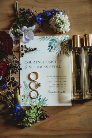 Wedding invitation flat lay for Courtney Umstot and Nicholas Stoll with gold rings, perfume bottles, and colorful florals.
