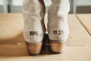 Close-up of white cowboy boots with MRS. and 11.21 wedding date stamped on heels, with engagement ring displayed between them.