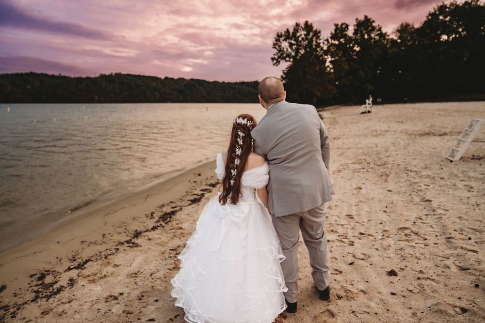 Bride and groom stand on sandy beach at sunset, gazing at calm lake with purple sky and trees.