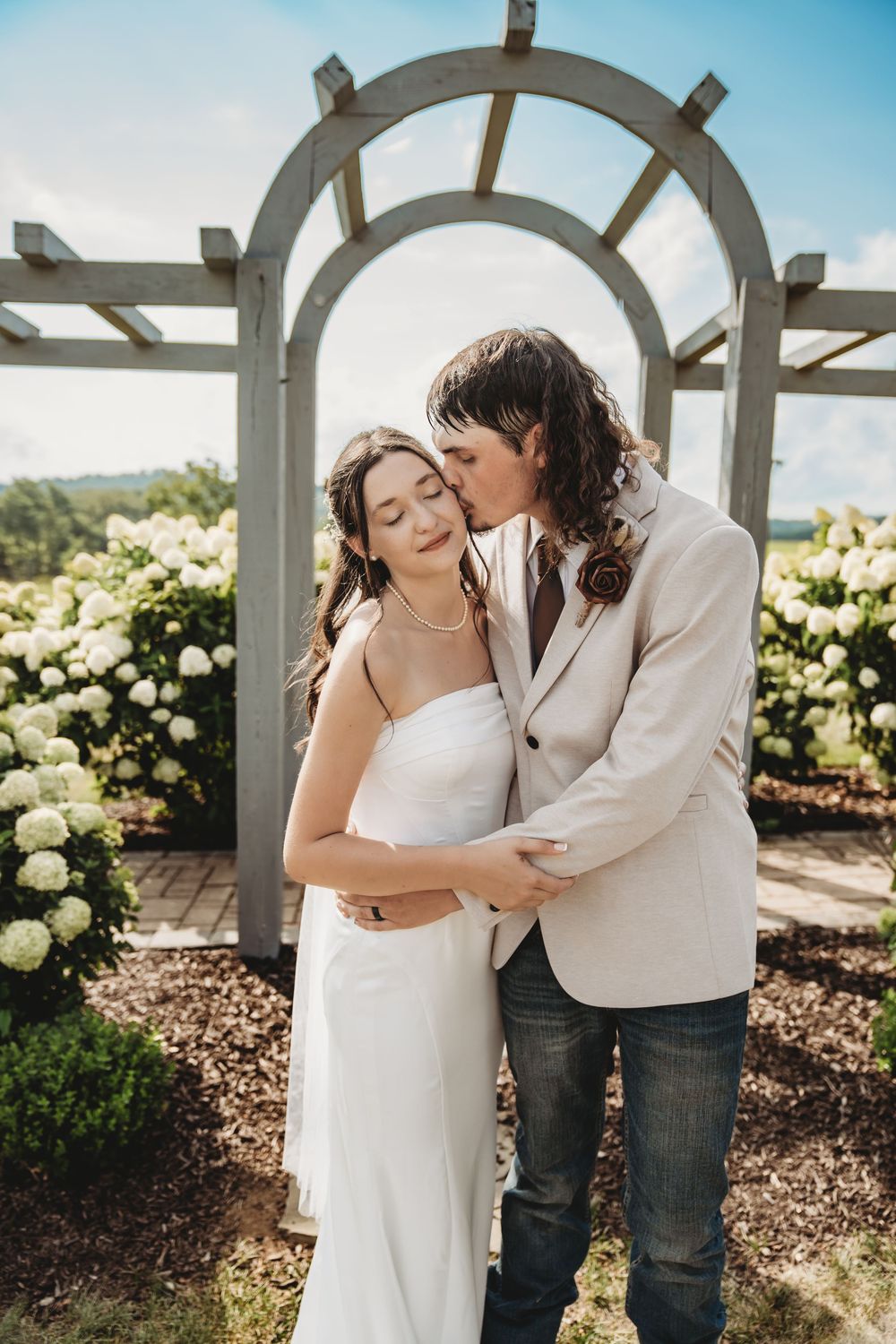 Bride and groom share a tender kiss under a white wooden arch surrounded by blooming hydrangeas on a sunny wedding day.