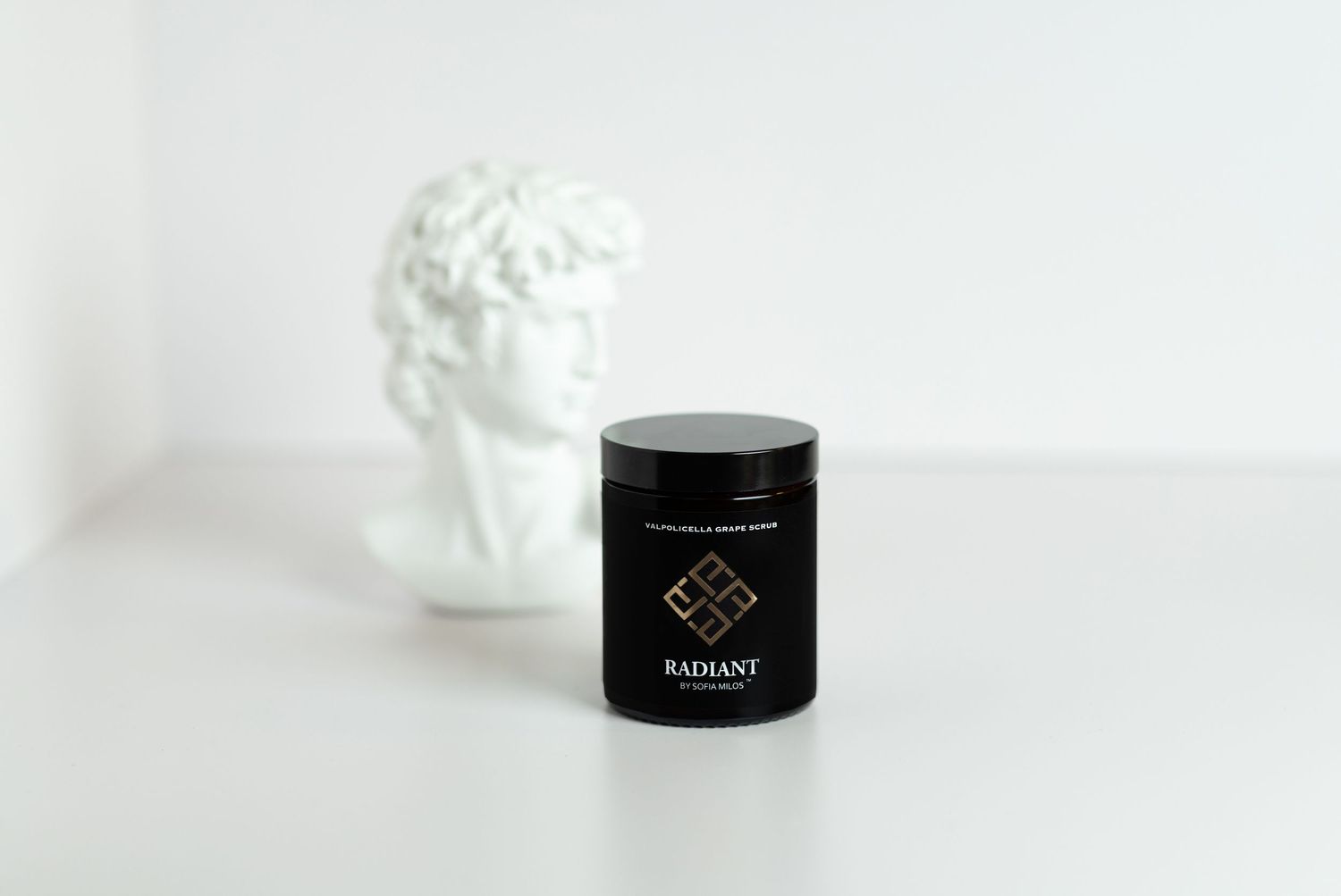 Minimalist product photography of black cosmetic jar with white bust sculpture against white background.