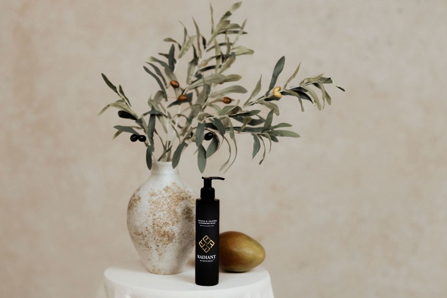 Elegant arrangement of olive branches in vase with black cosmetic bottle and fruit on white surface.