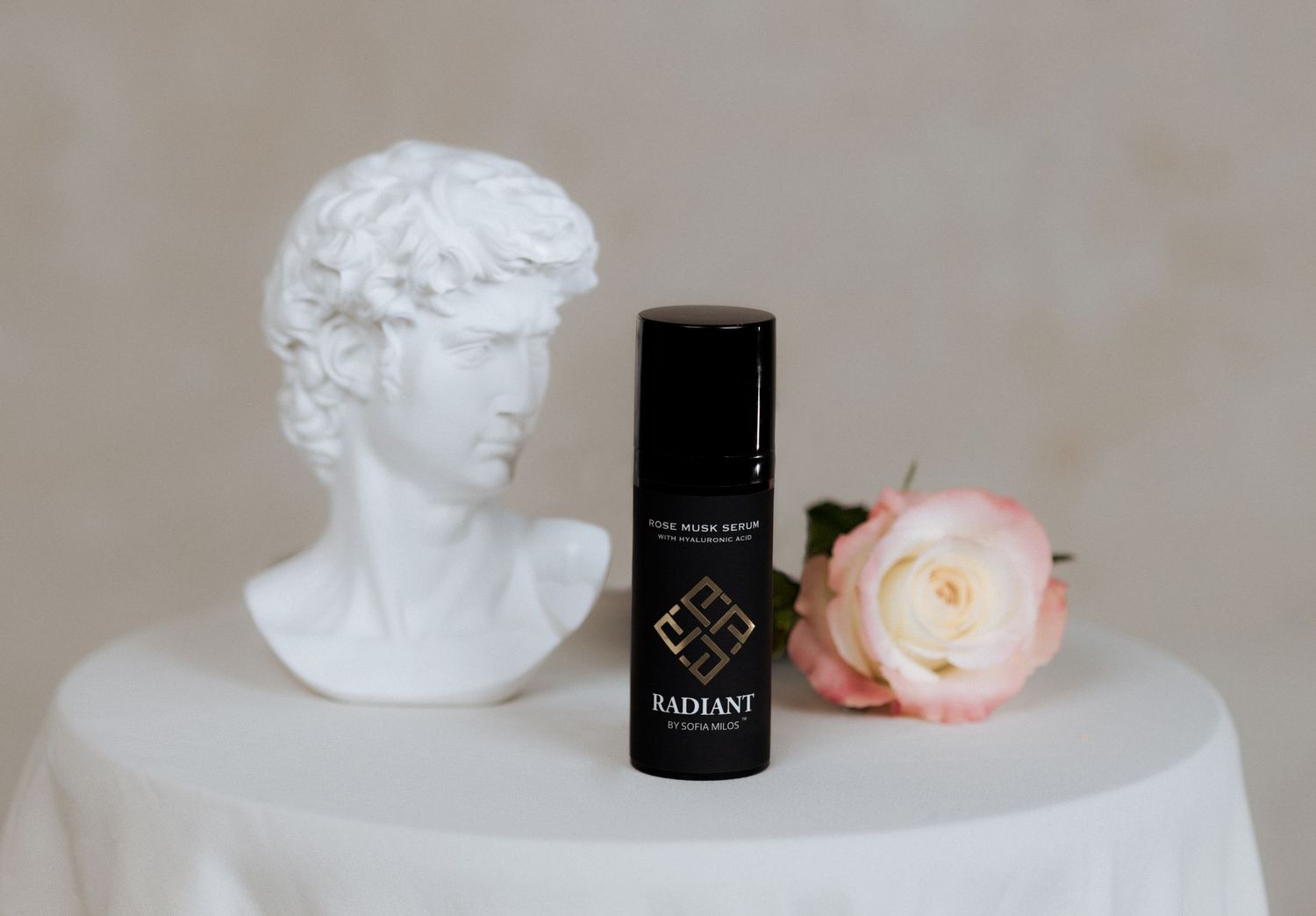 Black cosmetic product bottle displayed with classical white bust sculpture and pink rose on light surface.