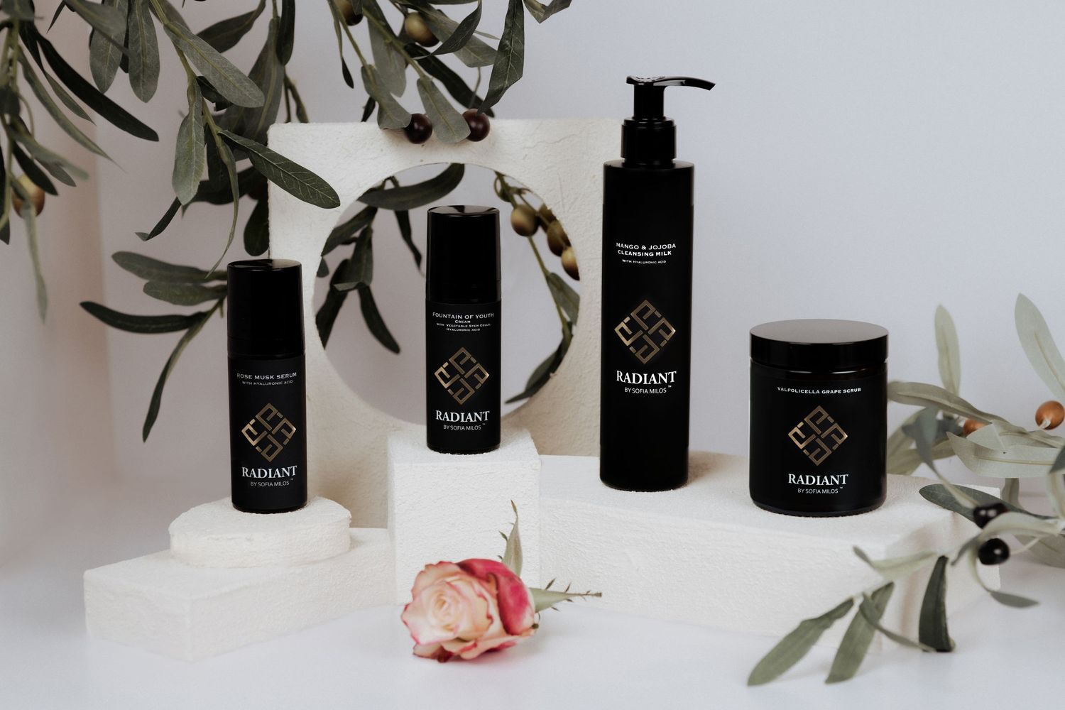 Collection of black cosmetic bottles and jars styled with olive branches and pink flower.