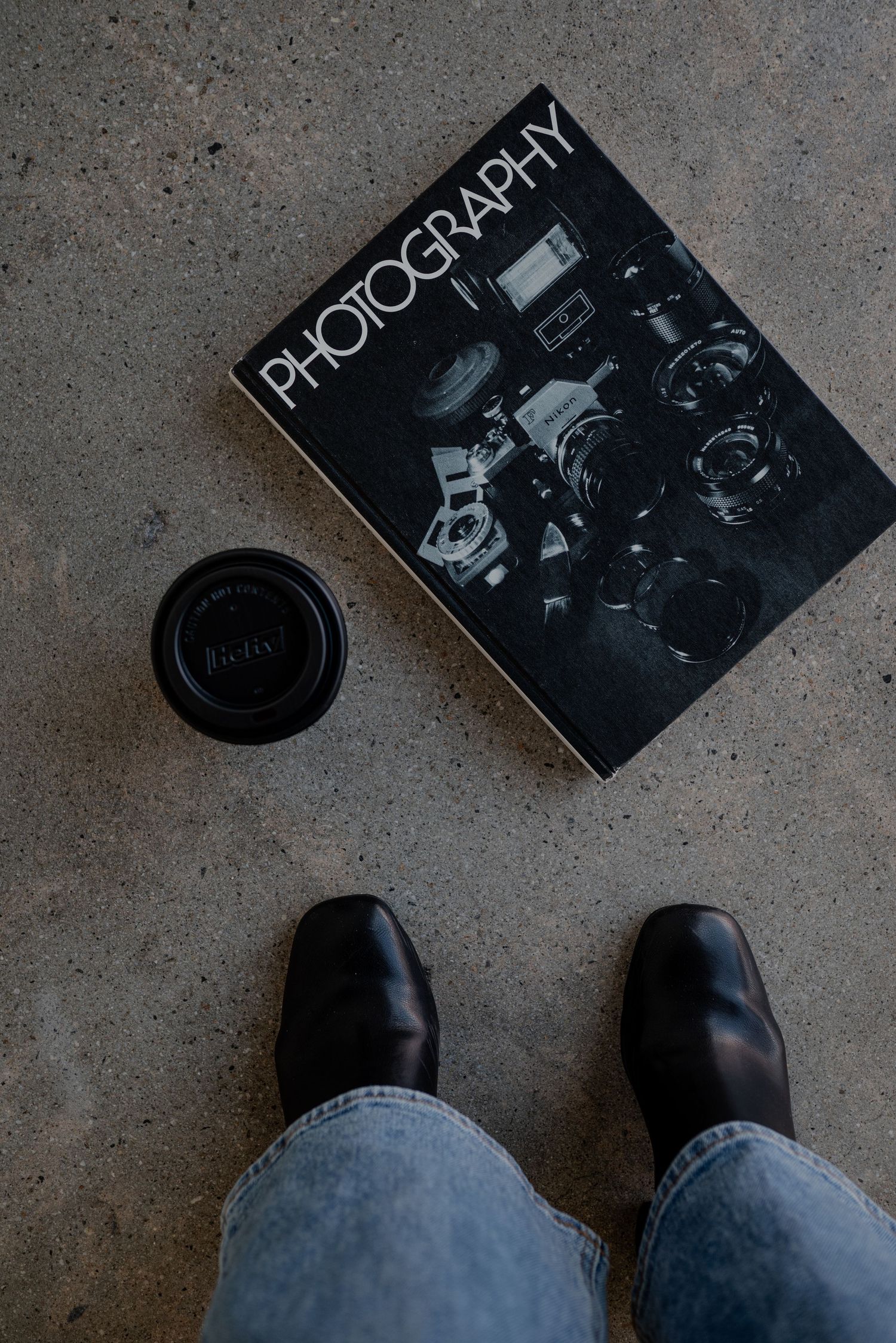 Overhead view of photography book and lens cap on concrete floor with feet in frame.